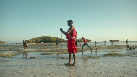 guerrero masai de pie en la playa con lugareños caminando en segundo plano en un día ventoso en watamu, kenia