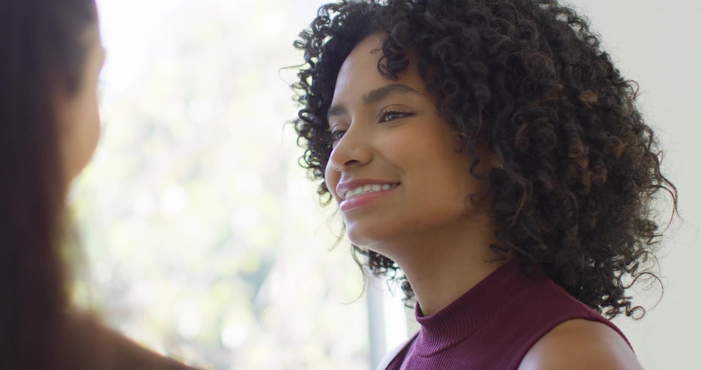 Woman listening and smiling at off-frame speaker by window in maroon top, copy space