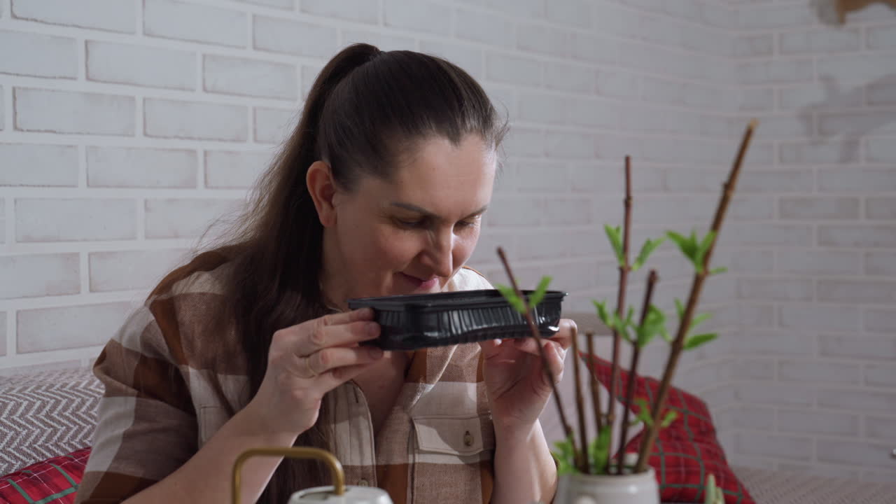 Woman lifts black plastic tray to sniff contents with subtle disapproval in bright indoor plant care setting on glass table near potted stems and watering can, closeup reaction moment