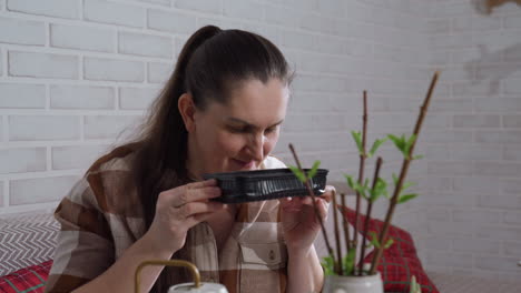 woman lifts black plastic tray to sniff contents with subtle disapproval in bright indoor plant care setting on glass table near potted stems and watering can, closeup reaction moment