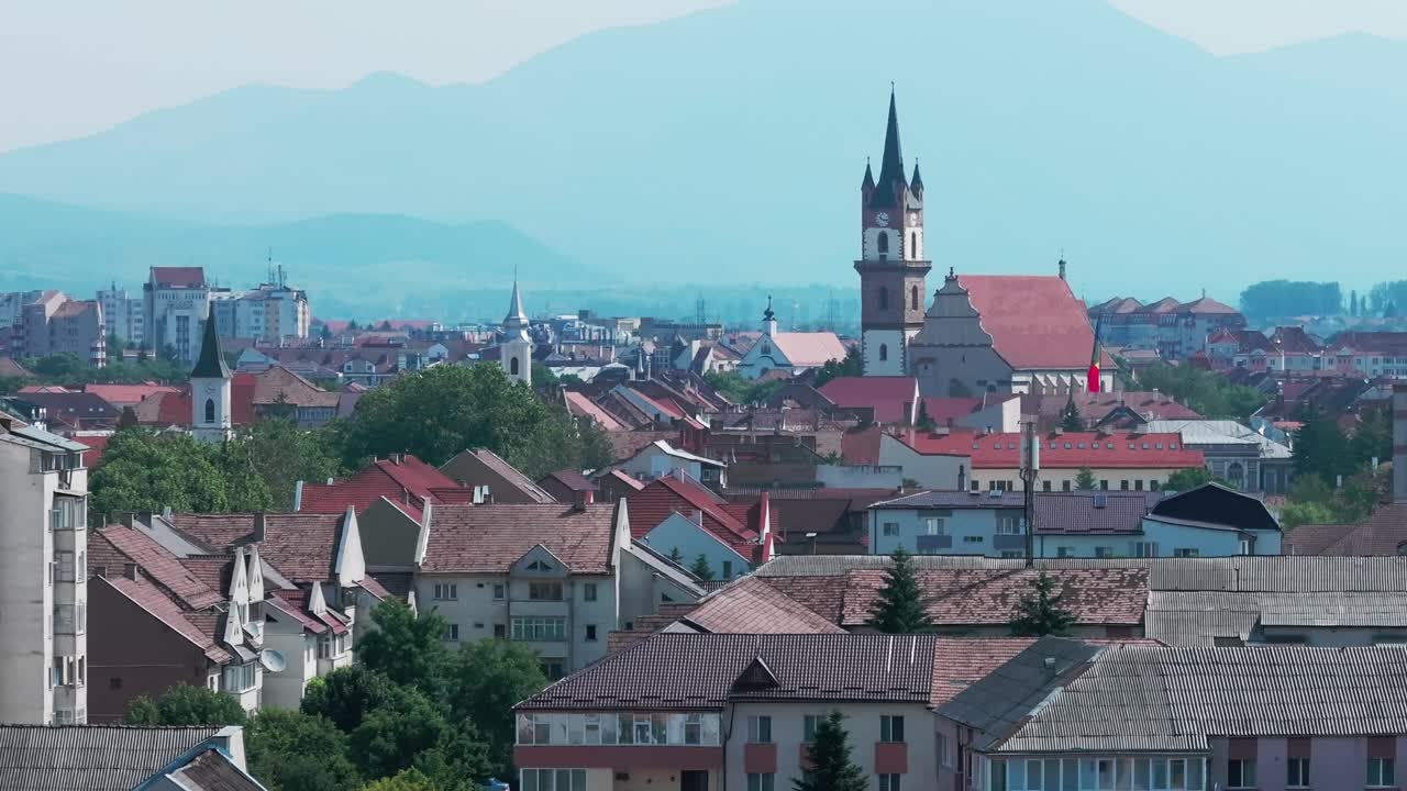 A aerial shot that focuses on the historic European cityscape of Bistrita, Romania. Tall tower of the Evangelical Church, surrounded by residential buildings and blue mountains in the distance