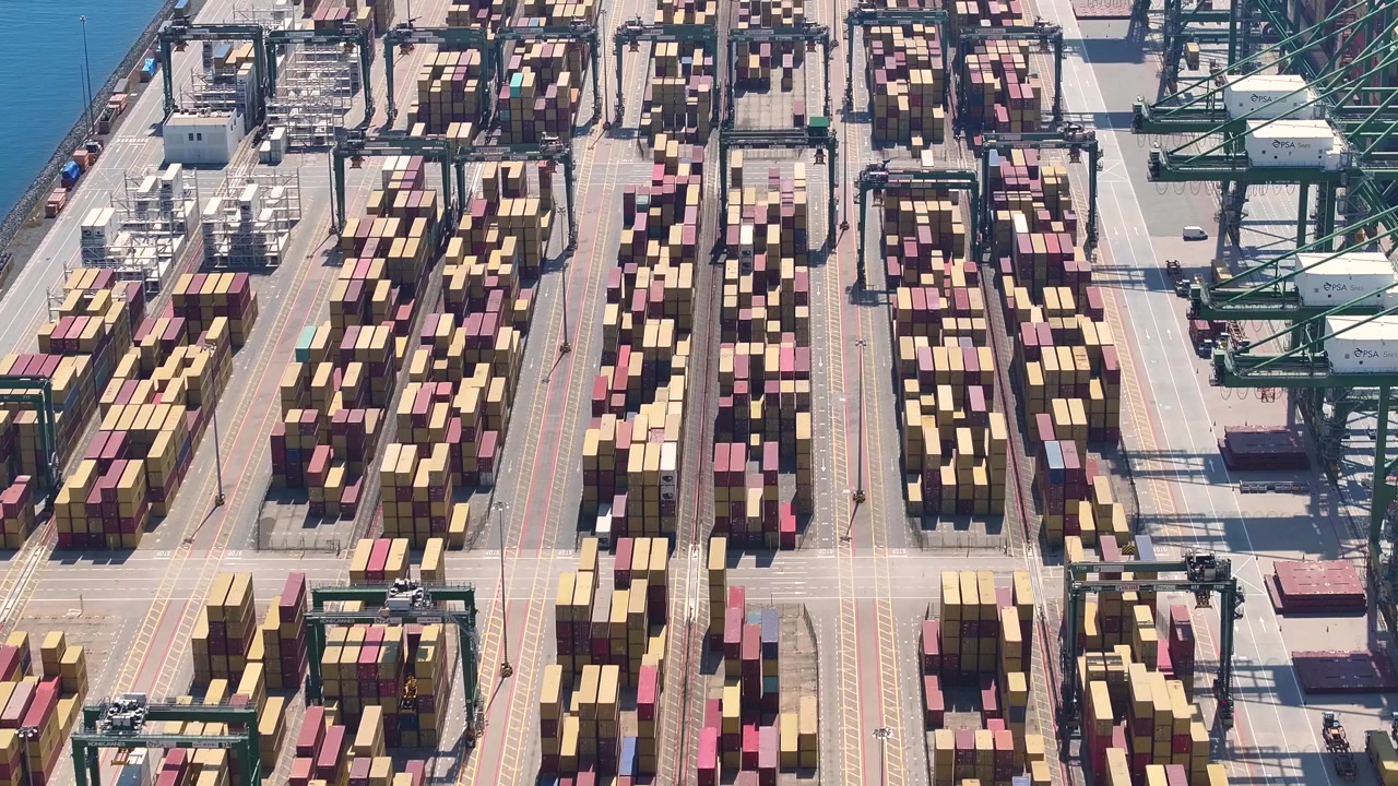 Containers are stacked at a busy port. Cranes move containers to and from ships. Delays are visible with congested cargo areas. Freight operations indicate high shipping volumes.