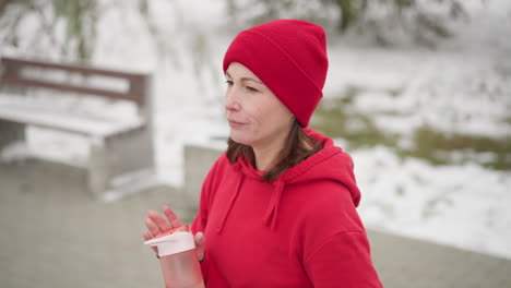 focused woman in red hoodie and beanie closes her pink water bottle outdoors, set against snowy background with greenery and blurred concrete bench