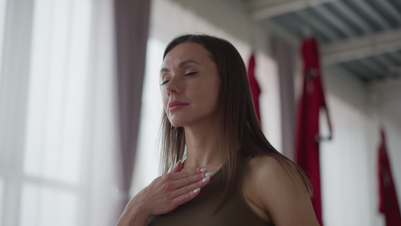 Fitness coach in sportswear stands in glass studio surrounded by fairy lights, places hands on chest in mindful meditative session, windows flood natural light into serene wellness space