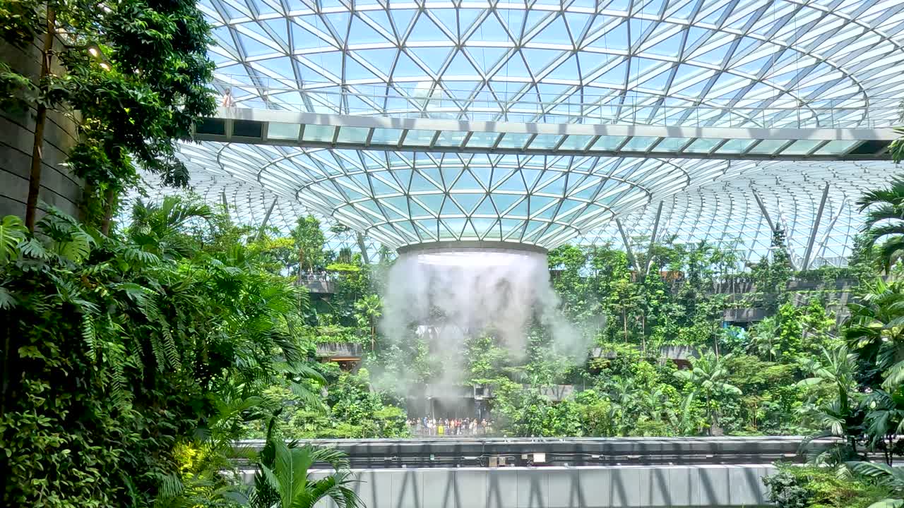 Large indoor waterfall flows amid tropical plants under geometric glass dome, bright daylight, static camera