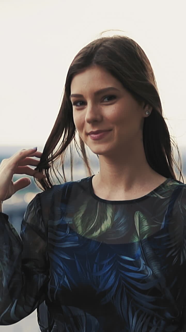 Coquettish young woman in elegant outfit with print decor touches hair adjusting near viewpoint handrail against large city slow motion closeup