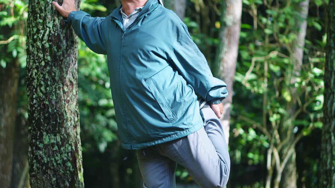Active senior asian man stretching legs while cling to tree at forest