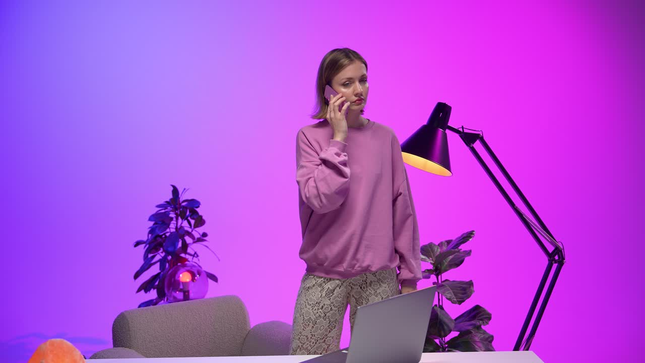 A young woman in a pink sweatshirt looks frustrated during a phone call while standing by a desk. The bold purple lighting, indoor plants, and casual workspace highlight modern emotions and tech use
