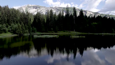 Panorámica-Aérea-Del-Bosque-De-Pinos-De-Los-Alpes-Franceses-Con-Nubes-En-El-Cielo-Azul-Sobre-Las-Rocas-De-Las-Montañas-Reflejadas-En-El-Lago-De-Las-Minas-De-Oro-[traducción:-Lago-De-Las-Minas-De-Oro]-Tour-De-Francia-Deportes-De-Vacaciones-Durante-El-Verano