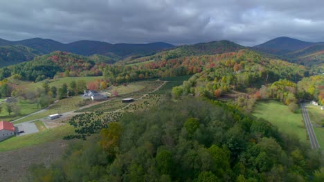 drone turns and glides over a mountainous valley with farmland and country homes below while dark storm clouds roll over and autumn leaves color the scene