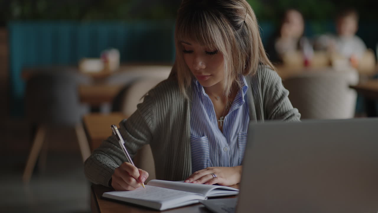 Asian woman working on her laptop and writing in his notebook sitting at a table. Working in coffee shop. woman looking to a laptop screen and making notes in her notebook. studying online.