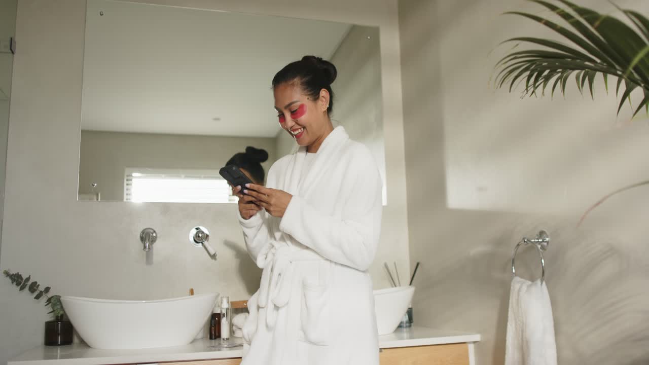 Asian mid adult female laughing at smartphone message and scrolling on bathroom sink, copy space