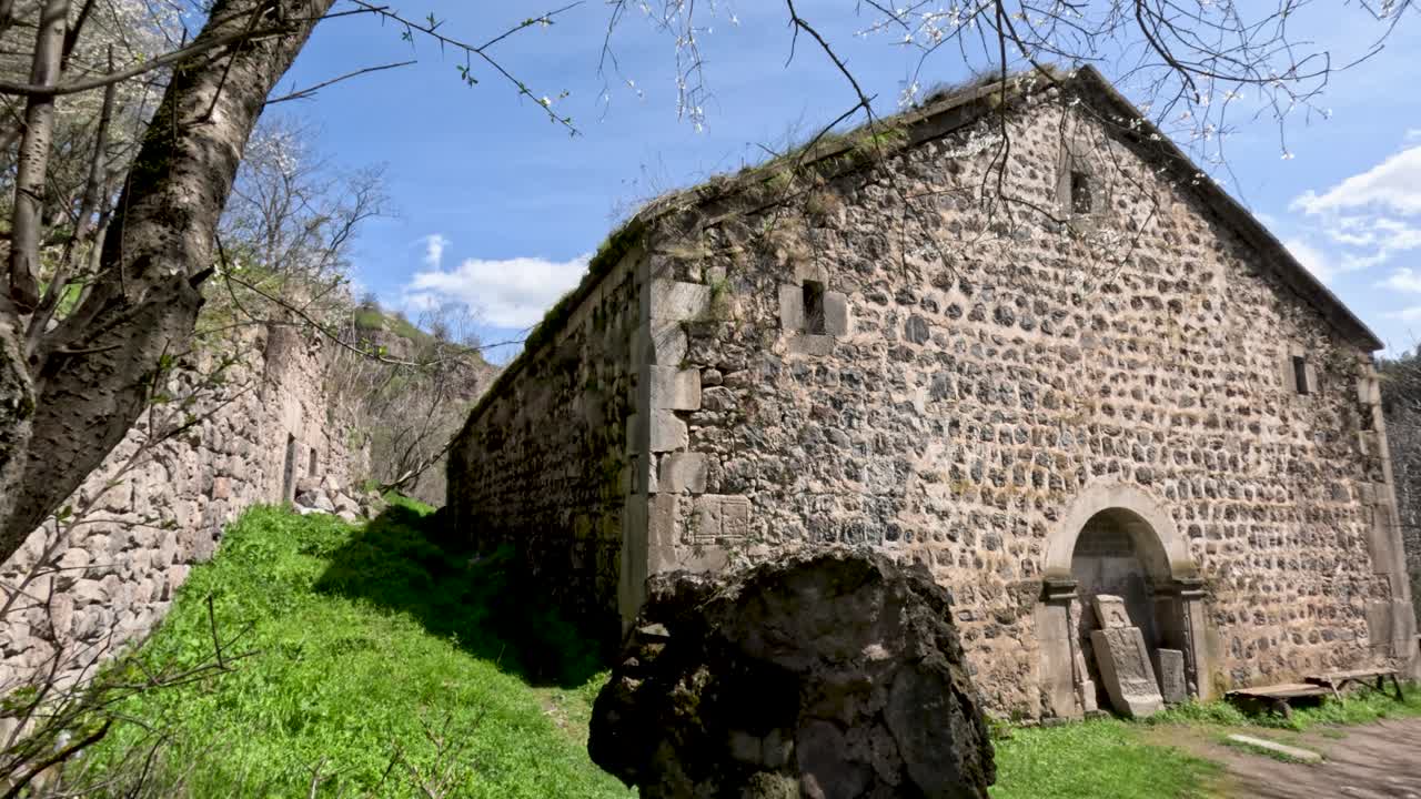 Due vecchi edifici in pietra si trovano in una verde valle. Il sole splende luminoso nel cielo blu. Alberi ed erba circondano l'area, aggiungendo alla bellezza naturale.