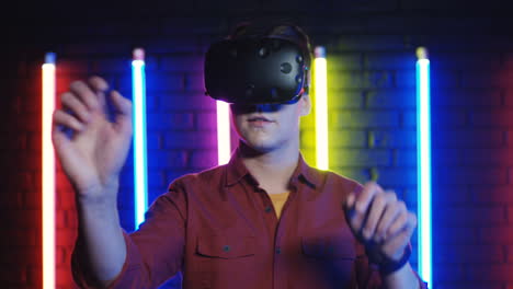 close up view of young man in the vr glasses moving his hands in the air in a room with colorful neon lamps on the wall