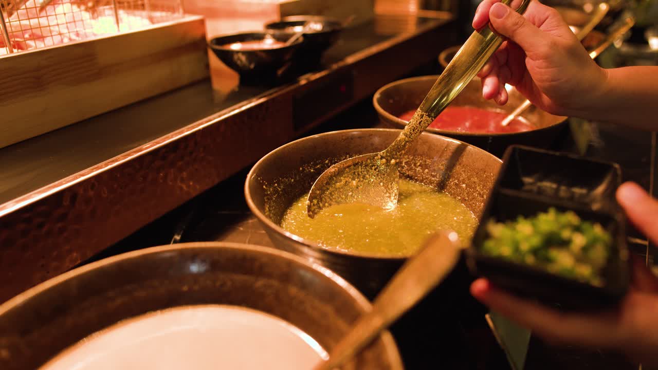 Hand ladles spicy broth into bowl at buffet, warm lighting, close-up, shallow depth of field