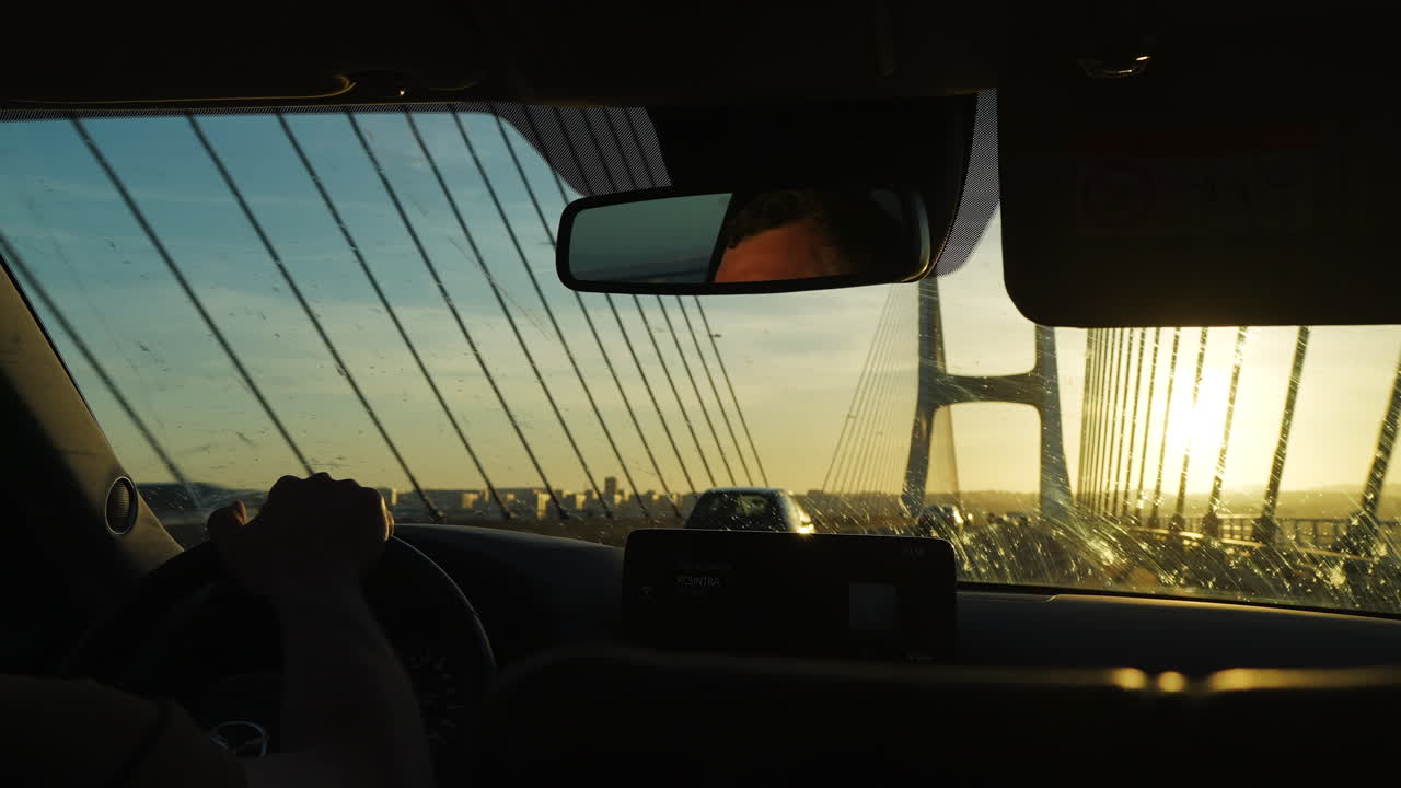 Driving through golden hour, car interior reveals Lisbon's iconic Vasco da Gama Bridge crossing the urban landscape, illuminating a scenic journey with warm sunset glow and reflections dancing across the windshield