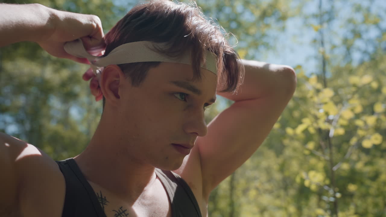 Close up view of soldier stretching white band with hands before tying on forehead while scanning forest surroundings, sweat and freckles visible on skin, sunlight filtering through trees