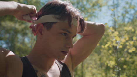 close up view of soldier stretching white band with hands before tying on forehead while scanning forest surroundings, sweat and freckles visible on skin, sunlight filtering through trees