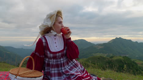 Junges-Blondes-Weibliches-Model-In-Traditioneller,-Altmodischer-Renaissance-Kleidung,-Das-In-Zeitlupe-Einen-Roten-Apfel-Isst,-Während-Es-Allein-In-Der-Berglandschaft-Sitzt