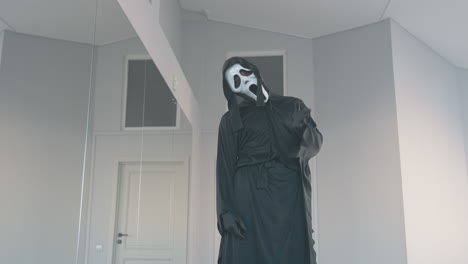 masked figure in flowing black costume steps toward camera in minimalist studio, hooded cloak and eerie white mask creating dramatic silhouette, subtle mirror reflection