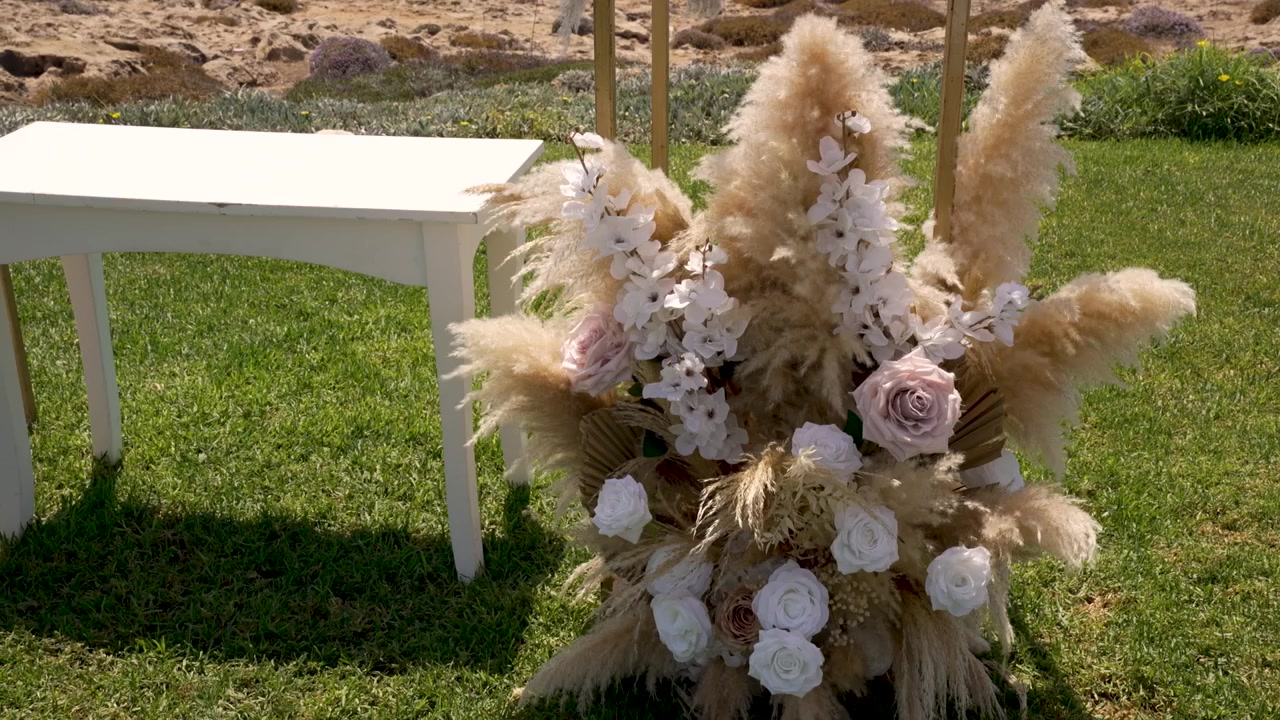Um arranjo de flores feito com rosas brancas e capim-dos-pampas está ao lado de uma mesa em um casamento perto da praia em um local ensolarado. O cenário oferece uma bela vista da natureza.