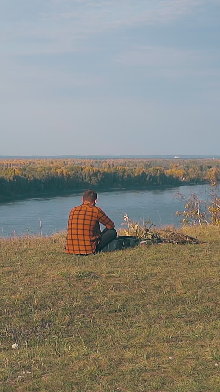 lonely guy in orange shirt with hangover sits at bonfire by blue tent in early morning slow motion backside view