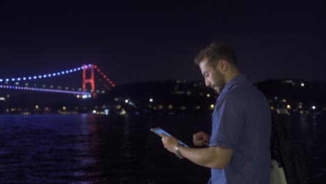 hombre usando tableta cerca del puente iluminado en el estrecho.