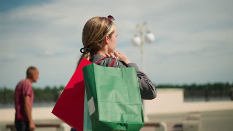 young woman walks outdoors carrying three colorful shopping bags on her shoulder, with a blurred background featuring a man walking by, the setting includes a pavement, street lamps