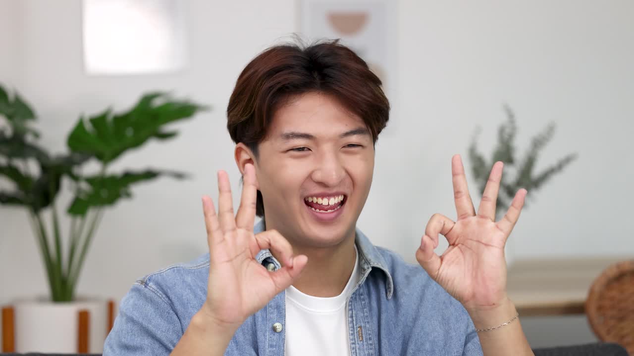 ชายหนุ่มชาวเอเชียที่มีความสุขยิ้มและแสดงสัญลักษณ์โอเคสองครั้งในห้องที่สว่างสดใส