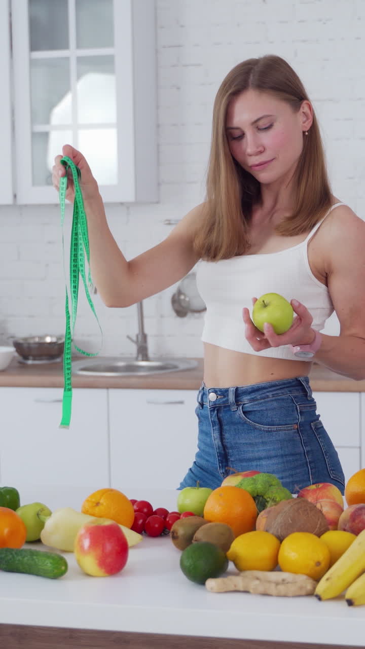 Sporty woman standing near fruits. Happy young woman with muscular body standing in the kitchen. Vertical video
