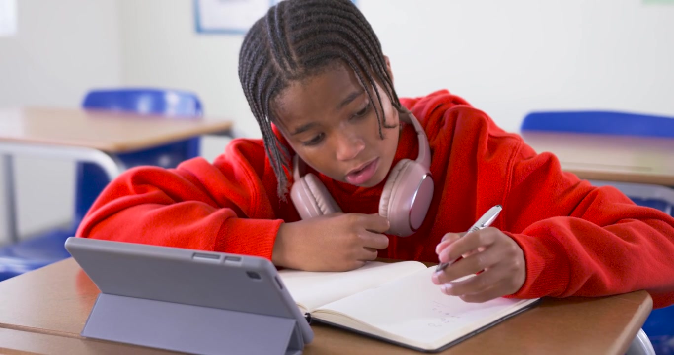African teen glancing at tablet, picking pen at school desk and writing in notebook while studying