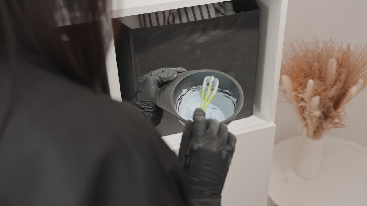 gloved hands mixing resin with sprig in small bowl artisan workspace on white shelf with black storage bins and dried pampas grass, minimal neutral interior, closeup of pouring and embedding