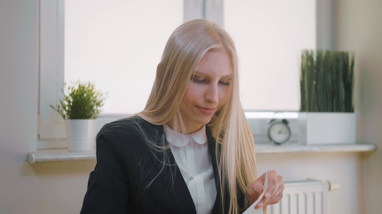 아름다운 금발 여성 슈트 책상에 앉아 노트북에 펜으로 글을 쓰고, 페이지를 뒤집고 노트북에서 종이를 꺼내고 있습니다.