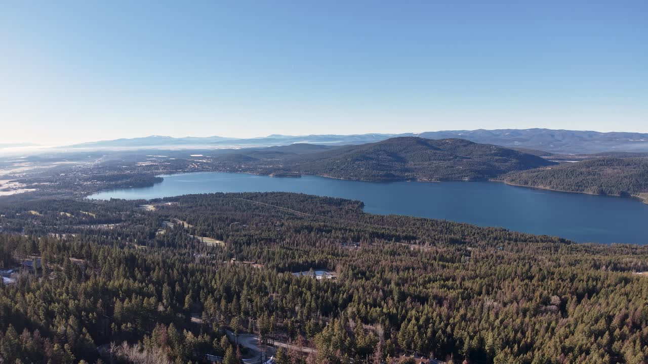 Letecký pohled dronem na široké horské jezero v Montaně, obklopené hustými jehličnatými lesy a zvlněnými kopci pod jasně modrou oblohou