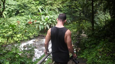 un hombre joven y en forma vestido de negro baja los escalones y las escaleras hacia un río en la exuberante y verde jungla de bali