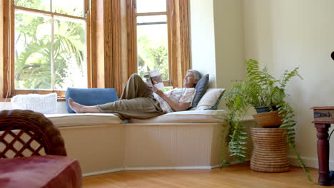 Happy-senior-biracial-woman-on-couch-reading-book-at-window-at-home,-slow-motion