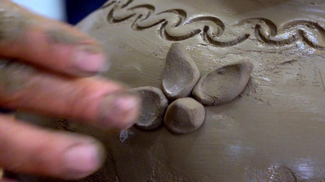 Primo piano di mani artigiane che modellano dettagli decorativi in argilla su una superficie di ceramica fatta a mano durante il processo tradizionale di lavorazione della ceramica.