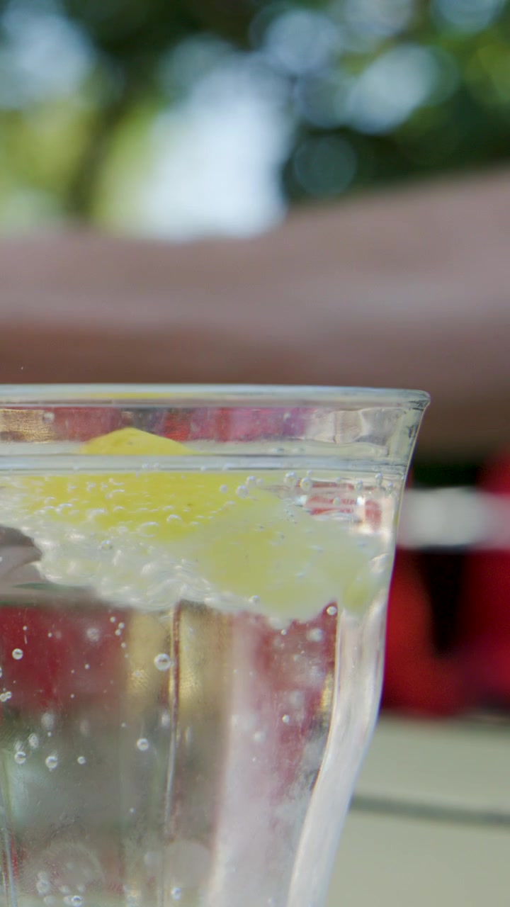 Hand stirs lemon slice in sparkling soda glass outdoors, natural daylight, shallow depth of field