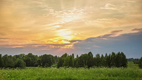 Scenic-view-of-orange-and-golden-sunset-behind-dark-clouds-in-timelapse-over-the-green-grasslands-and-trees