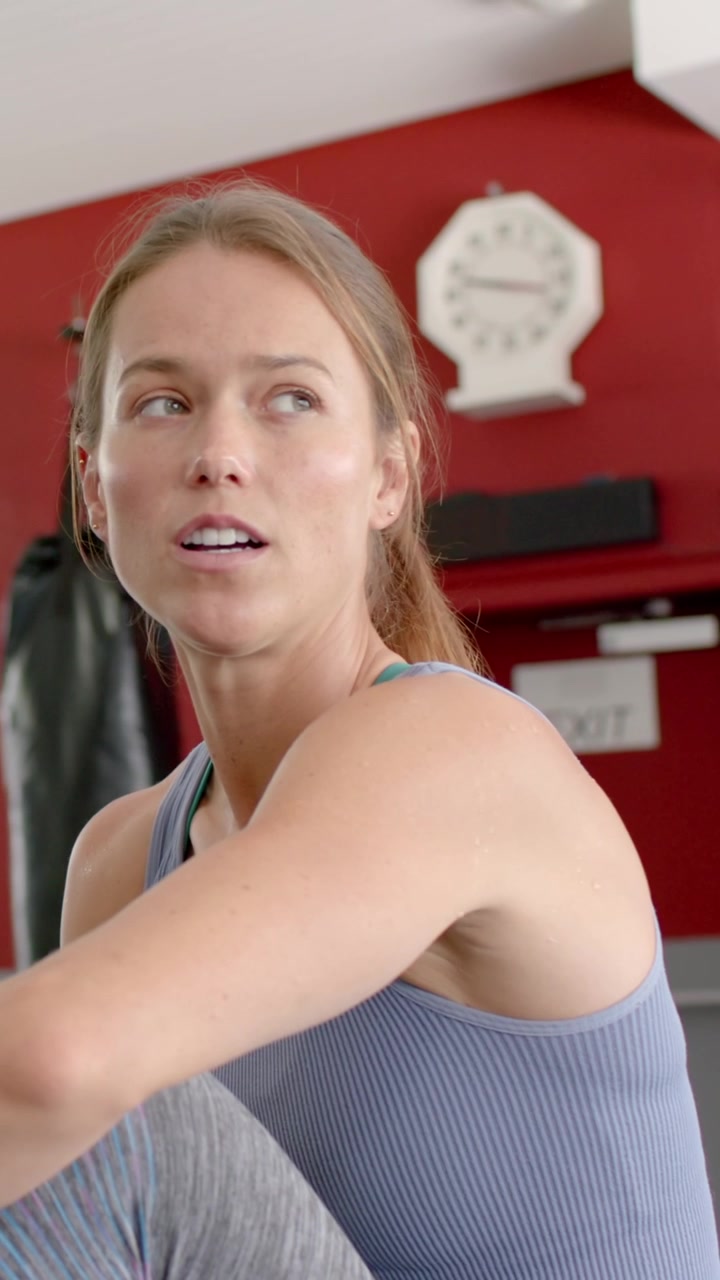 Vertical video: Woman in workout gear sitting in gym resting between sets, turning toward EXIT sign