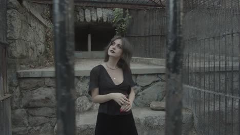 Young-girl-posing-in-a-black-dress-trapped-in-a-cell,-view-through-prison-bars