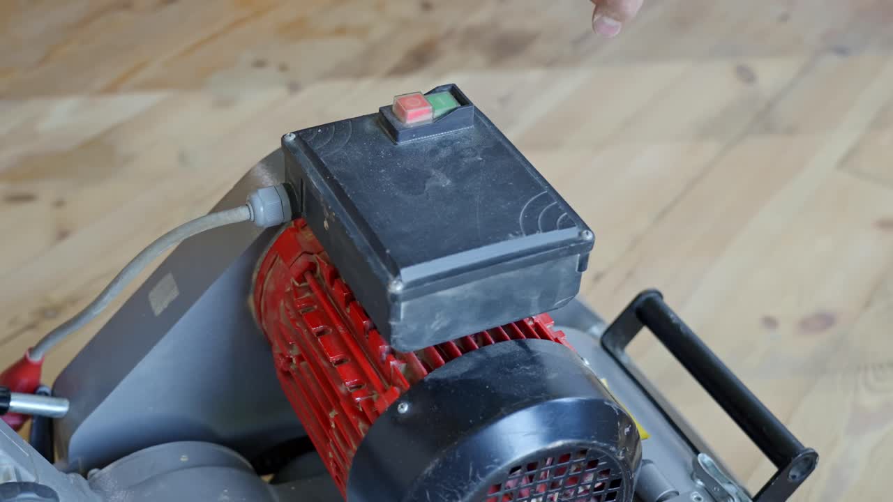 Close-up shows hand pressing the power button on industrial wood floor sander with red motor, situated on a pine wood floor, illustrating preparation for sanding in home renovation project static shot