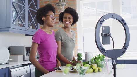 happy african american mother and daughter preparing healthy drink, making video using smartphone