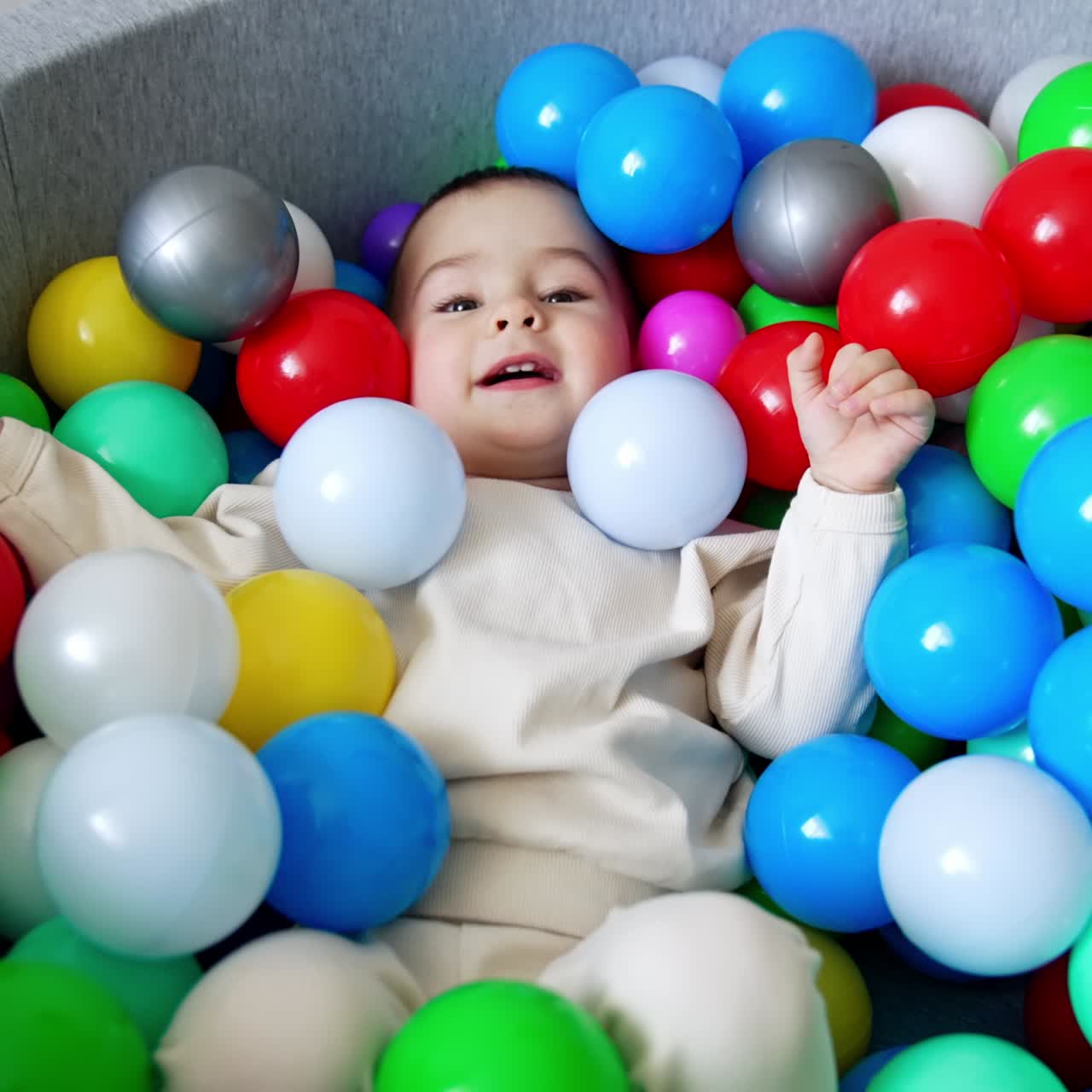 Adorable toddler sits in a dry pool. Cute boy leans back and lies in the multiple balls smiling to the camera