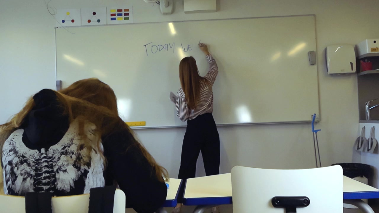 Giovane insegnante che scrive su una lavagna durante una lezione in un'aula moderna, con una studentessa che osserva attentamente dal suo banco in primo piano, pronta a prendere appunti e imparare