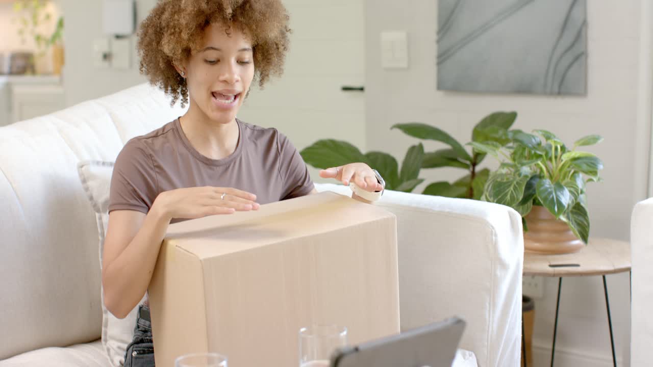 African American woman and pal speaking holding opening box on sofa tilting toward tablet to show
