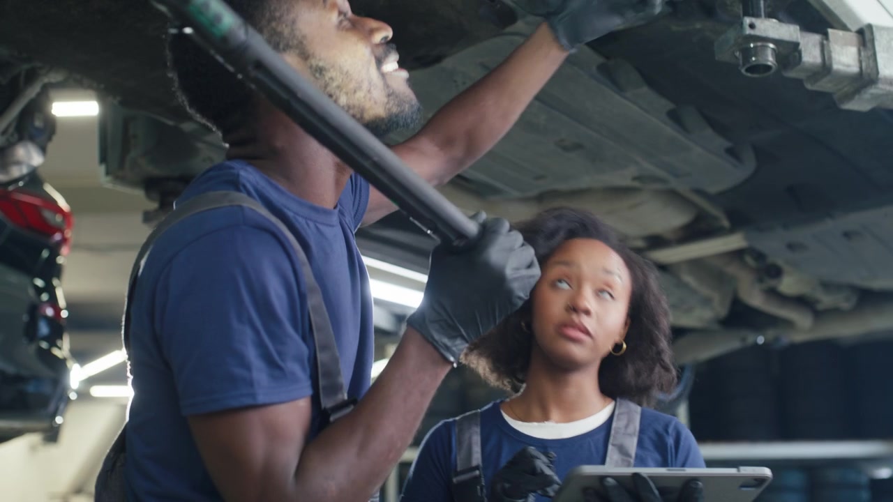 Two african american mechanics a man and a woman inspecting a vehicles undercarriage with a torch