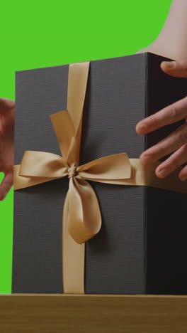 vertical video of man picking up present in gift wrapped box decorated with ribbon on table shot against green screen