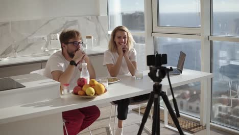 Young-happy-couple-blogger-talking-to-camera.-Having-breakfast-while-record-video-chat-on-camera.-speaking-on-camera-home-skype-technology-cheerful-people-discussion-blog-webcam,-online-video