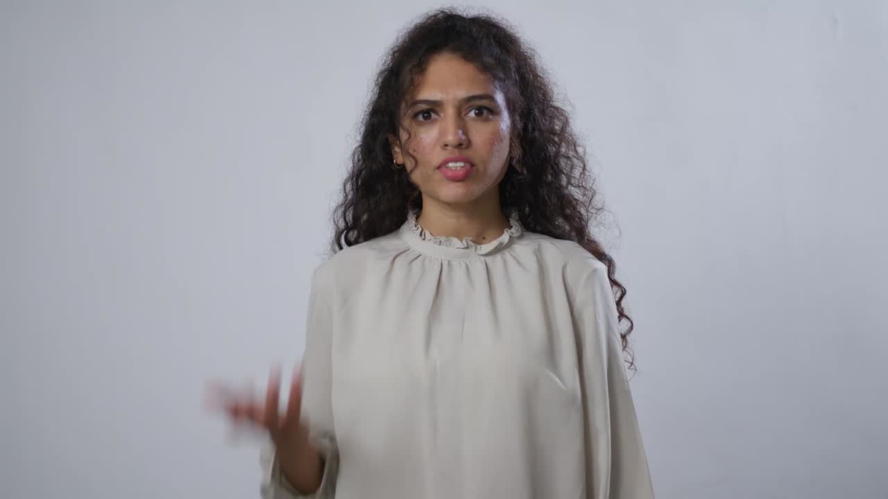 A South Asian woman with curly hair in a beige blouse, showing quiet disapproval or disappointment